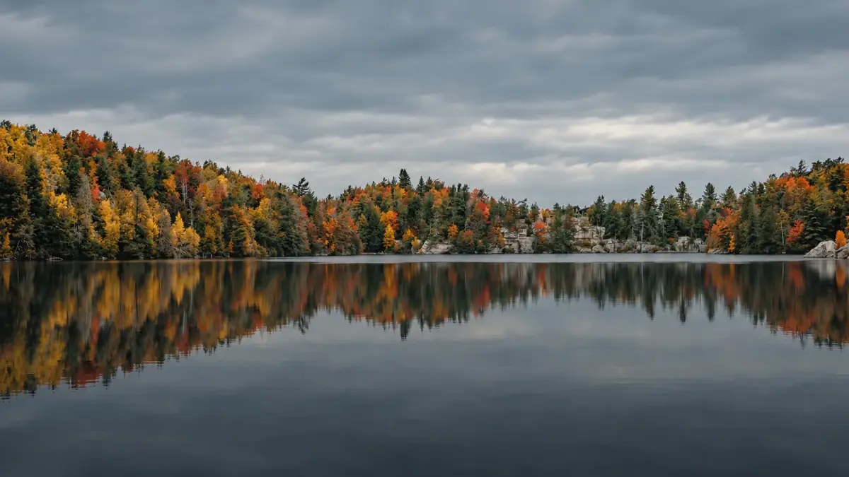 An autumn lake under a grey sky, its shoreline of red, orange and gold trees reflected in still water